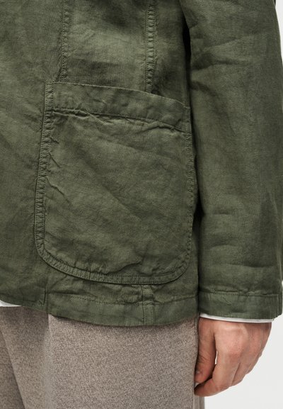 Κοντινό πλάνο τσέπης και μανικιού πράσινου λινό σακακιού πάνω από γκρί υφασμάτινο παντελόνι, με το χέρι να ακουμπά δίπλα στο σώμα.