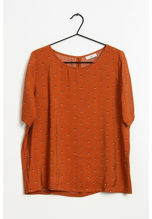 Bluzka w kolorze rdzawego pomarańczu z krótkim rękawem, z małym czarno-białym wzorem w kropki, okrągły dekolt, zawieszona na czarnym drucianym wieszaku na białym tle.