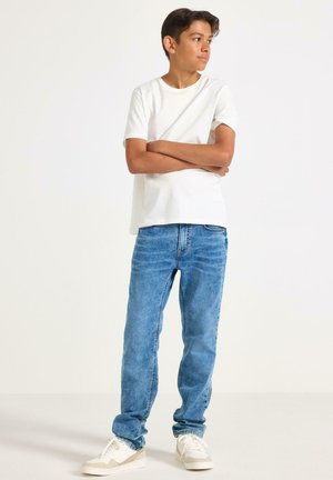 Ragazzo in piedi con le braccia incrociate, indossa una maglietta bianca, jeans blu e sneakers bianche, guarda a destra su uno sfondo bianco uniforme.
