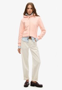 Joven mujer vistiendo una sudadera rosa con cremallera, camisa azul claro, pantalones crema, cinturón marrón y mocasines marrones oscuros, de pie con una mano en el bolsillo.