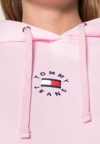 Hoodie rosa chiaro realizzato in misto cotone. Presenta il logo ricamato "TOMMY JEANS" con un design a rettangolo blu, bianco e rosso. Chiusura con cordino.