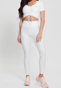 Žena v texturovaném bílém crop topu s nařasením a vysokých legínách s béžovými a růžovými teniskami, stojící na jednobarevném pozadí.