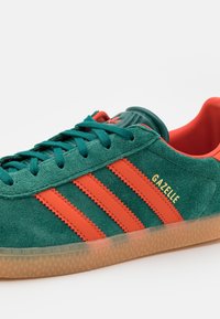 Green suede sneaker with orange leather stripes and golden text. Features a rubber sole and textured accents. Laces are teal.