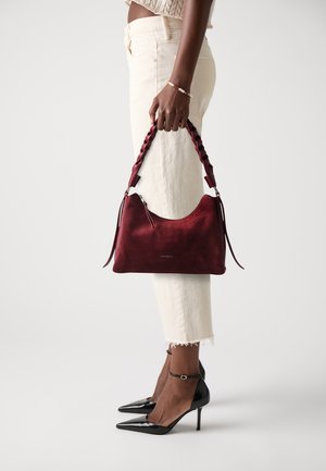 A person holds a red suede handbag with a braided strap, wearing white cropped pants and black high heels, set against a plain background.