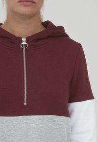 Haut à capuche coloré en maroon et gris, avec une fermeture éclair en argent, doté d'une capuche avec cordon de serrage et d'accents blancs sur les manches. Texture de tissu douce.