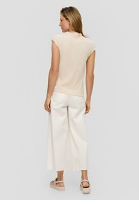 Crème, getextureerde mouwloze top met een ruffle halslijn, gecombineerd met brede witte broeken en lichtbeige plateau sandalen.