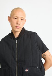 Črn brezrokavnik z zadrgo spredaj in rebrastim ovratnikom. Ima dva sprednja žepa in majhno logotipno nalepko; izdelan je iz teksturirane tkanine.