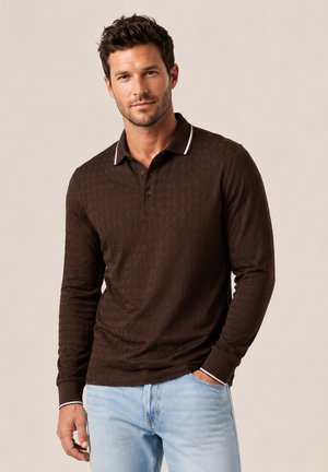 Hombre con una camisa polo de manga larga marrón oscuro y vaqueros azul claro, de pie con una mano en el bolsillo contra un fondo neutro.