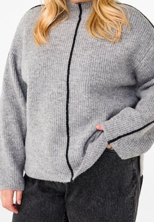 Femme portant un pull gris en tricot avec une bande verticale noire, partiellement rentré dans un jean noir, main dans la poche.