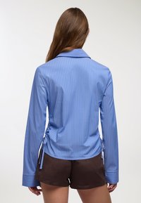Donna con lunghi capelli castani che indossa una camicia a righe blu con maniche lunghe e nodi laterali, insieme a pantaloni corti marroni scuri, vista da dietro.
