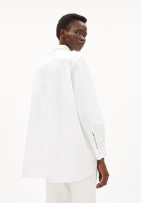 White long-sleeve shirt with a loose fit, featuring a rounded hem, button cuffs, and green accents peeking from the sleeves. Smooth texture.