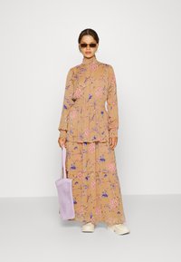 Vestido longo e em camadas, feito de um tecido castanho claro com padrões florais em rosa e azul. Combinado com uma bolsa de malha lavanda e ténis brancos.