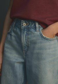 Ljusblå denimjeans med en blekt finish, med en framknäppning, bälteshällor och sidofickor. Materialet verkar mjukt med en sliten textur.