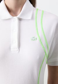 Polo bianco con tessuto a trama, caratterizzato da un logo ricamato verde e due linee di accento verdi lungo la spalla e il lato.