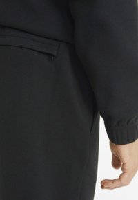 Svart hettegenser med glidelåslomme på siden, ribbestrikkede mansjetter og en myk tekstur, som fremhever et enkelt og funksjonelt design.