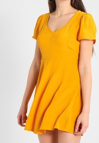 Gul kort klänning med v-ringning, puffärmar, åtsittande bodice och utsvängd kjol. Tillverkad av lätt tyg med diskret textur.