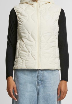 Gilet sans manches matelassé couleur crème avec fermeture éclair à l'avant, porté sur un t-shirt noir à manches longues et un jean bleu clair.