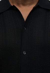 Gros plan d'une personne portant une chemise en maille texturée noire à boutons avec col, montrant la partie supérieure de la poitrine et du cou.