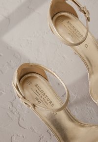 Sandales à talons métalliques dorés avec brides chevillères, texture lisse et semelle intérieure de marque. Fabriquées au Brésil, avec des détails complexes sur les brides.