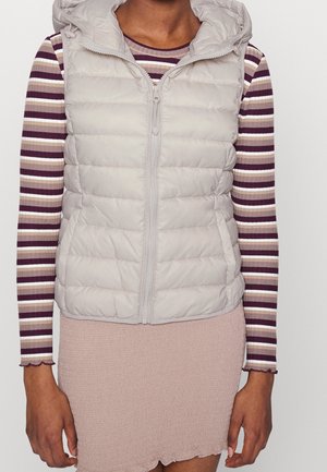 Woman wearing a light gray quilted hooded vest over a long-sleeve striped shirt and textured pale pink skirt.