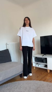 Bela prevelika majica s črnim besedilom, kombinirana s črnimi rebrastimi hlačami. Okoli je siva kavč, televizija in radiator.