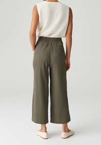 Kalhoty s širokými nohavicemi z texturované olivově zelené tkaniny, s vysokým pasem, zadní kapsou a uvolněným střihem, v kombinaci s bílým tílkem bez rukávů.