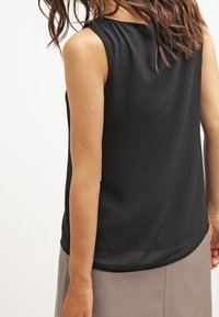 Kobieta w czarnej bluzce bez rękawów i beżowej spódnicy, pokazana z tyłu z luźnymi falującymi włosami.