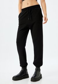 Schwarze Sweatpants mit elastischem Bund, schmal zulaufenden Bündchen und Seitentaschen; weicher Jersey-Stoff; kombiniert mit schwarzen Knöchelschuhen.