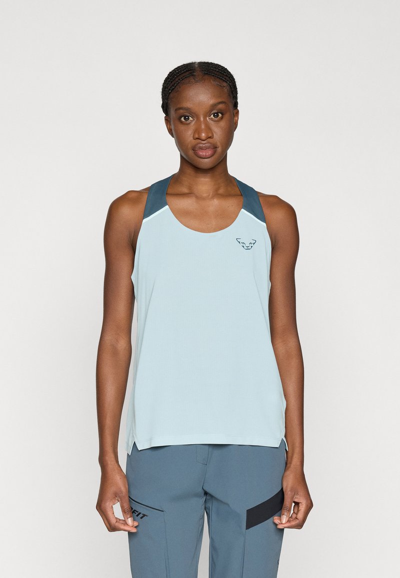 Γυναίκα ντυμένη με ανοιχτό γαλάζιο αμάνικο αθλητικό τοπ και μπλε-γκρι παντελόνι, στέκεται με το πρόσωπο στραμμένο μπροστά σε μια απλή υπόβαθρο.