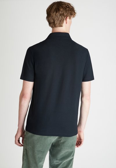 Camisa polo negra de manga corta con cuello clásico, textura suave y dobladillo recto, vista desde atrás. Combinada con pantalones de pana verdes.