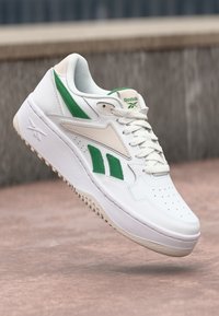 Biele tenisky s zelenými akcentmi, vyrobené z hladkej kože, s perforovanou špičkou, polstrovanou podrážkou a viditeľným logom Reebok na jazyku.