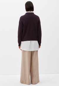 Personne se tenant de dos, portant un pullover à côtes violet foncé sur une chemise blanche et un pantalon large beige, sur un fond uni.