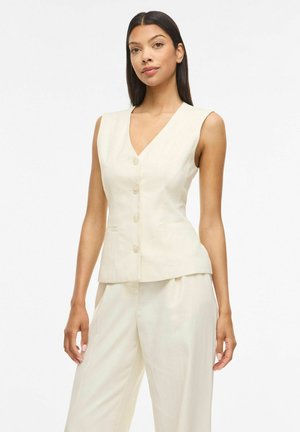 Mujer con chaleco sin mangas color crema con botones y pantalones sueltos de talle alto a juego, sobre un fondo blanco liso.