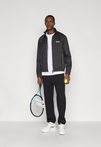 Μαύρο ελαφρύ τζάκετ με φερμουάρ και ψηλό γιακά, λευκό λογότυπο, σε συνδυασμό με λευκό T-shirt και μαύρη φόρμα, κρατώντας ρακέτα και μπάλα τένις.