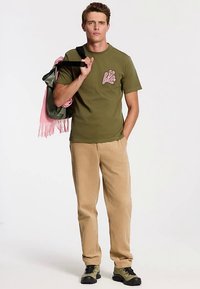 Olivno zelena majica s rožnatim grafičnim dizajnom, bež kordurove hlače, rožnati šal premeščen čez ramo in črna obutev.