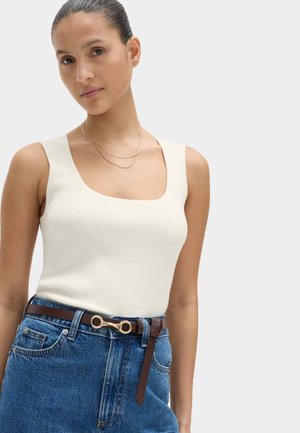 Femme portant un haut blanc sans manches à encolure carrée, un jean bleu avec une ceinture en cuir marron dotée d'une boucle dorée, et un collier doré superposé.