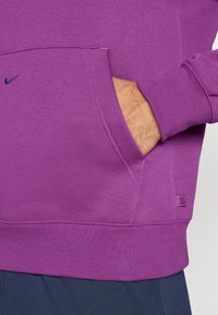 Lilla Nike sweatshirt med kengurulomme, ribbede manchetter og kant. Inkluderer en lille logoaccent på brystet. Blød stofstruktur.