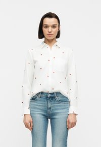 Νέα γυναίκα με κοντά σκούρα μαλλιά, ντυμένη με λευκό πουκάμισο με μικρά κόκκινα μοτίβα καρδιάς και ανοιχτόχρωμα μπλε τζιν, στέκεται μπροστά σε λευκό φόντο.