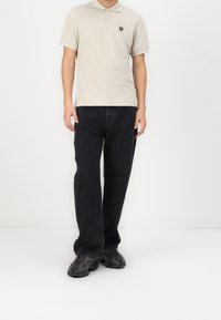 Polo shirt beige clair à manches courtes avec un petit logo sombre sur la poitrine, associé à un jean noir à coupe décontractée et des baskets noires texturées.