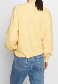 Pull en maille jaune à coupe décontractée, épaules tombantes et ourlet côtelé. Porté avec un jean en denim bleu taille haute.
