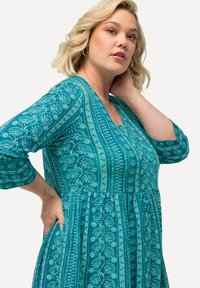 Vestido floral en tonos teal con mangas tres cuartos, que presenta un escote redondeado, cintura fruncida y un intrincado diseño en todo el tejido.