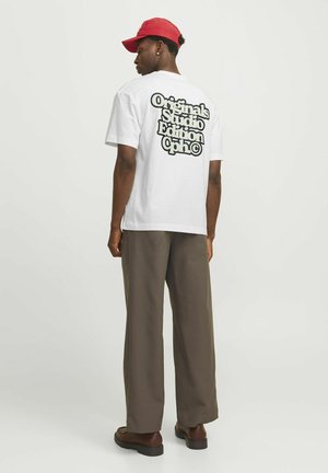 Tricou alb din bumbac, având pe spate un text mare, de culoare neagră, care spune "Originals Studio Edition Cph." Asortat cu pantaloni maro lejeri și o pălărie roșie.