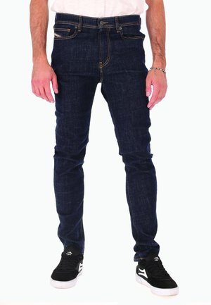 Man in een donkerblauwe slim-fit jeans, zwart-witte sneakers en een wit shirt, staand met ontspannen handen naast zich.