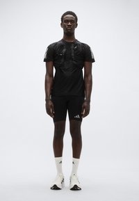 Μαύρο αθλητικό πουκάμισο με ιμάντα, μαύρο στενό σορτς, λευκές κάλτσες και λευκά αθλητικά παπούτσια. Χαρακτηρίζεται από διακριτικό λογότυπο και εφαρμοστή σχεδίαση.