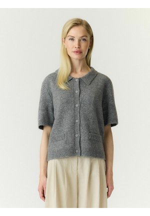 Femme aux cheveux blonds raides portant un cardigan gris à manches courtes boutonné et un pantalon beige à jambes larges sur un fond clair.