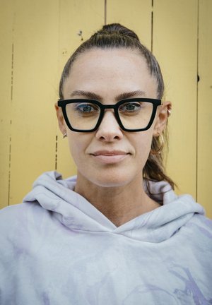 Czarne, duże okulary z przezroczystymi szkłami, jasnofioletowa bluza z kapturem z wzorem oraz kolczyk-koło na uchu na żółtym drewnianym tle.