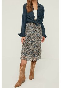 Žena v džínové košili svázané v pase, bílem crop topu, květinové midi sukni a hnědých semišových kovbojských botách.