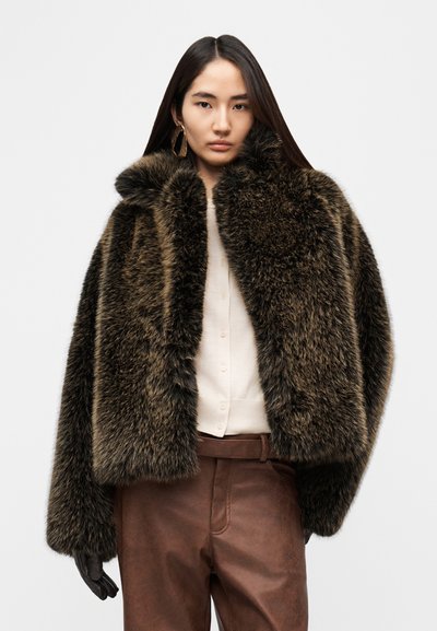 Veste en fausse fourrure marron foncé avec un col montant, des manches larges et une finition texturée. Portée sur une chemise claire et un pantalon marron.