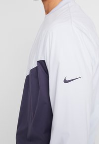 Nike bunda s bílými a tmavě fialovými panely, hladkým syntetickým povrchem, vroubkovaným límcem a nenápadným logem na rukávu.