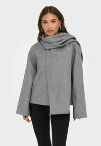 Grå ullblandad kappa med ett draperat, överdimensionerat krage, vida ärmar, asymmetrisk framknäppning och mjuk textur. Stylerad med svarta byxor.